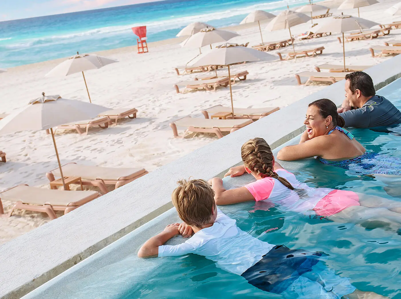 Family on vacation in an infinity pool overlooking the beach with chairs and beach umbrellas.